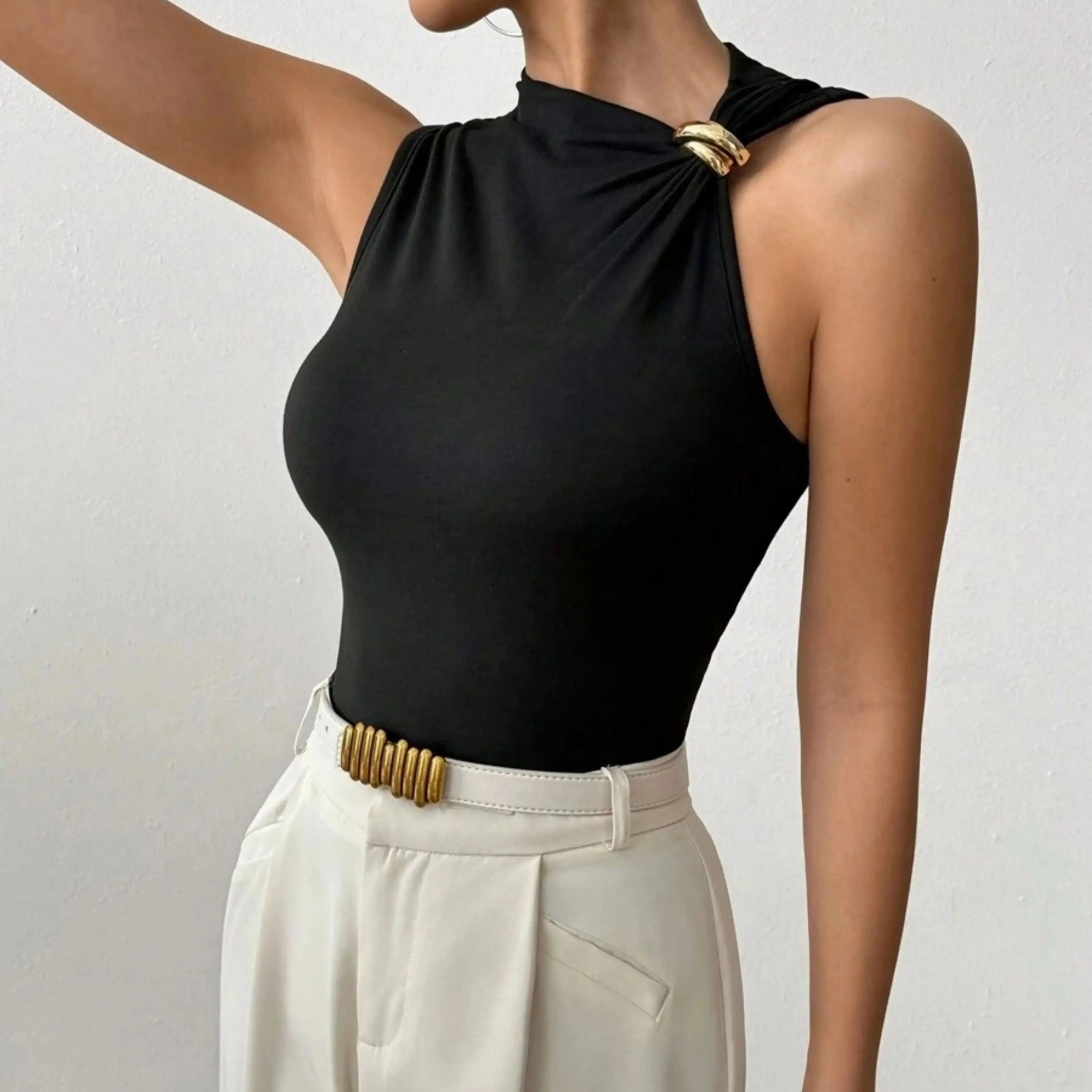Person wearing a black sleeveless top with gold accents and white pants on a plain background